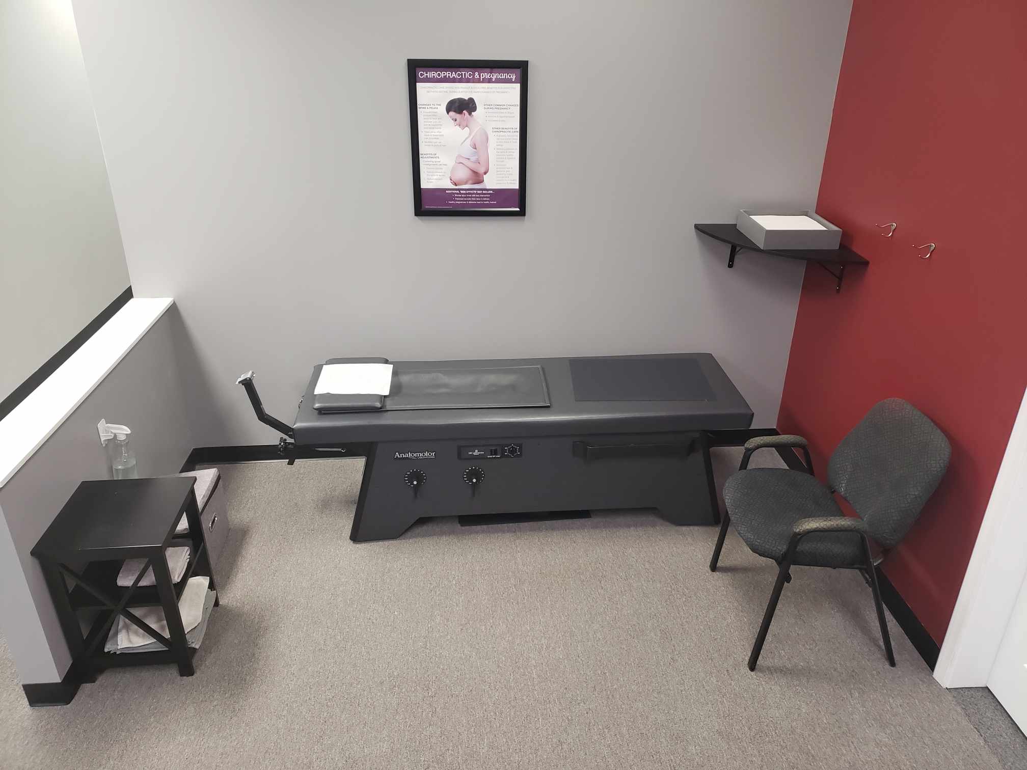 Treatment room with a gray roller massage table, a chair and a red accent wall